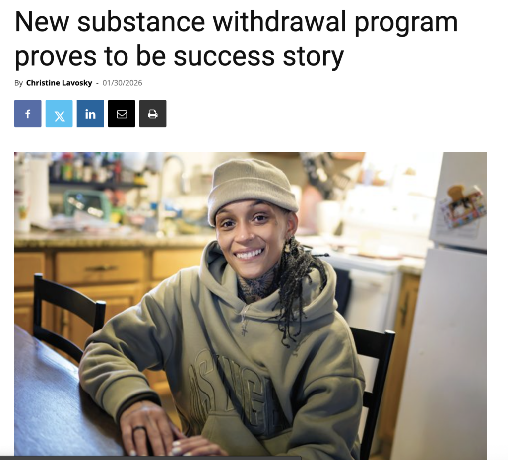 Mercedes Faust sits at her kitchen table smiling. She wears a gray sweatshirt and a gray beanie.